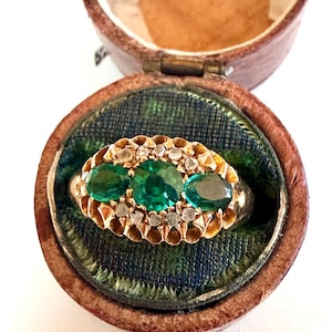 May include: A vintage gold ring with three emeralds and small diamonds, set in a decorative band. The ring is displayed in a brown leather jewelry box with a green and blue patterned interior, creating a classic and elegant presentation.
