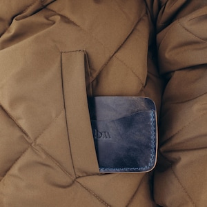 Puede incluir: Una cartera de cuero azul con costuras azules es parcialmente visible en el bolsillo de una chaqueta acolchada marrón. La cartera tiene un aspecto desgastado y las letras "DM" están grabadas en la parte delantera.