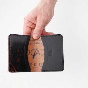 Op de afbeelding: Een zwart en bruin leren kaarthouder met een bi-fold ontwerp. De binnenkant heeft een beige gekleurd materiaal met de tekst "LL CORDO" en "OGAD" erop gedrukt. De kaarthouder wordt opengehouden en toont de binnenkant.