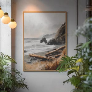May include: A framed landscape art print depicts a coastal scene with a cloudy sky, ocean waves, and driftwood on the beach. The artwork is in a wooden frame and hangs on a gray wall. The overall color palette is muted with shades of gray, brown, and beige.