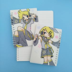 May include: White spiral-bound notebooks with cartoon illustrations of a character with yellow hair, wearing a gray and yellow outfit. The notebooks have lined pages and are decorated with pink and blue stars.