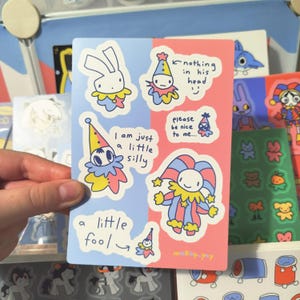 May include: A sheet of colorful stickers featuring cartoon jesters and a bunny. The stickers have a blue and pink background with text that reads "I am just a little silly", "nothing in his head", and "please be nice to me".