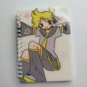 May include: A notebook with a spiral binding, showcasing a vibrant anime-style illustration of a character with yellow hair and a grey and yellow outfit. The cover is protected by a clear plastic overlay with pink star and flower details.