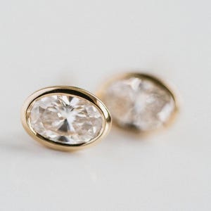 May include: A pair of gold stud earrings featuring oval-cut, clear gemstones. The gemstones are set in a gold bezel. The earrings are presented on a white backdrop, with one earring in focus and the other slightly blurred.