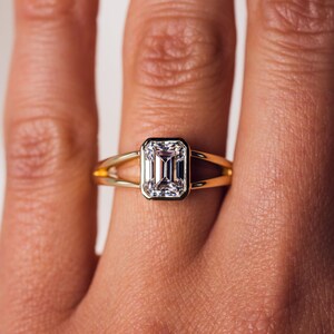 May include: A gold ring featuring a rectangular emerald-cut diamond. The diamond is set in a bezel setting with a black border. The ring has a split shank design.
