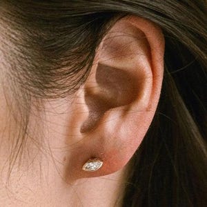 May include: Close-up of an ear with a gold stud earring. The earring features a marquise-cut diamond set in a gold bezel. The earring is small and delicate, adding a touch of sparkle.
