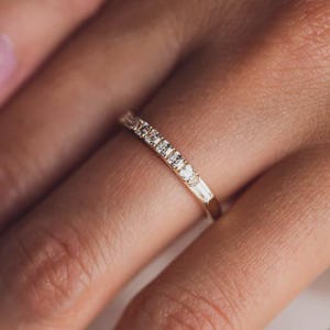 May include: A gold ring featuring a line of small, clear diamonds. The ring is worn on a finger, with the focus on the ring's details. The diamonds are rectangular and square-cut, and the band has a smooth, polished finish.