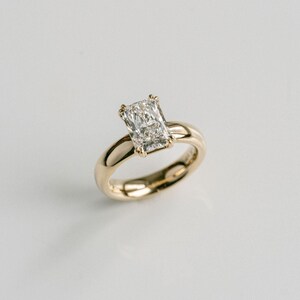 May include: A gold ring featuring a single rectangular radiant-cut clear gemstone set in a four-prong setting. The band is smooth, polished, and solid gold, showcasing a classic design with a high-shine finish that reflects light across the surface.