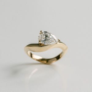Puede incluir: Un anillo de oro con un diamante en forma de pera. La banda del anillo tiene un diseño fluido y asimétrico. El diamante está engastado en una montura de garras. El anillo se muestra sobre una superficie blanca.