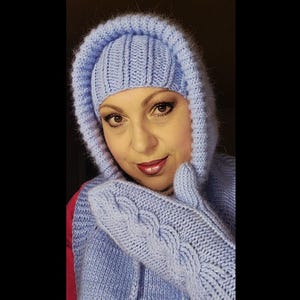 May include: A light blue knitted winter set, including a hooded hat, scarf, and mittens. The mittens feature a cable knit design. The set is made of a soft, textured yarn, perfect for cold weather. The hat has a ribbed design.