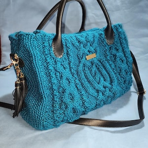 May include: Teal knitted handbag with a cable knit design. The bag features brown leather handles, a detachable shoulder strap, and a gold-tone metal tag. A matching tassel hangs from the side.