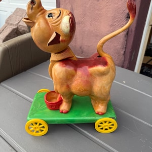 May include: Vintage pull-along toy in the shape of a pig. The pig is tan with red accents and is mounted on a green platform with yellow wheels. A small red bucket is attached to the platform.