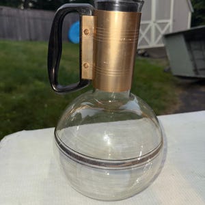 May include: A vintage glass decanter with a black stopper and handle. The decanter has a gold-colored metal band around the neck and a decorative band around the base. The handle is black and attached to the gold band. The decanter is sitting on a white surface.