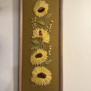 May include: Embroidered wall art featuring yellow sunflowers with brown centers and green leaves, set against a muted green background. The artwork is framed in a light brown wooden frame, creating a vintage aesthetic.