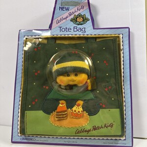 May include: A new Cabbage Patch Kids tote bag in its original packaging. The bag features a doll with a yellow and green hat and scarf. The bag is green with a design of two cupcakes on a plate. The packaging reads "NEW Cabbage Patch Kids Tote Bag."