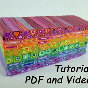 Può includere: Una pila colorata di canne in pasta polimerica con motivi geometrici in tonalità di rosa, arancio, giallo, verde, blu e viola. Le canne sono disposte in un motivo a strati, creando un design visivamente accattivante e intricato. L'immagine include il testo "Tutorial PDF e Video".