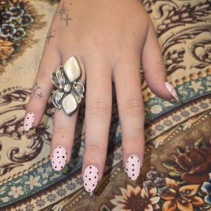 May include: A hand with a large silver ring featuring a flower design and two white stones. The nails are painted light pink with black polka dots. A black bracelet with white text is visible on the wrist.