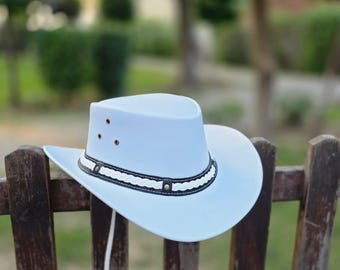 May include: A light blue cowboy hat with a wide brim and a decorative band. The band features a black and white woven design with small brass rivets. A white chin strap is attached. The hat is resting on a wooden fence.
