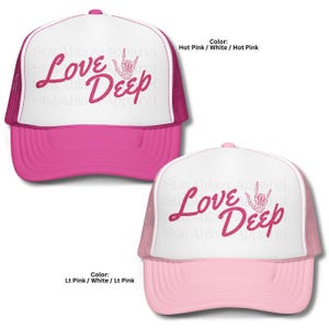 Może przedstawiać: Dwie czapki typu trucker z napisem "Love Deep" w różowym kolorze i grafiką gestu dłoni. Jedna czapka jest w kolorze intensywnego różu z białym przodem, a druga w kolorze jasnoróżowym z białym przodem. Czapki mają siatkowy tył.