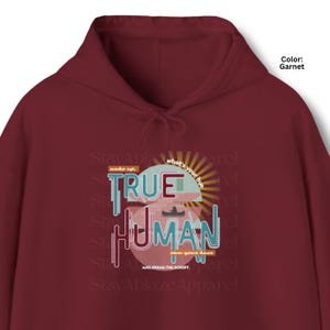 Op de afbeelding: Een granaatrode hoodie met een grafisch ontwerp met de tekst "TRUE HUMAN" in grote blauwe en witte letters. Het ontwerp bevat een zonnestraal, het silhouet van een kano en de zinnen "wake up," "what's your hue," en "own your hues."