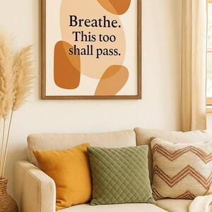 以下が含まれることがあります： 「Breathe. This too shall pass.」の文字が黒で書かれた額入りのウォールアートプリント。アートワークは、茶色とベージュの色合いの抽象的な形を特徴としています。ベージュのソファの上に、装飾的な枕が置かれています。