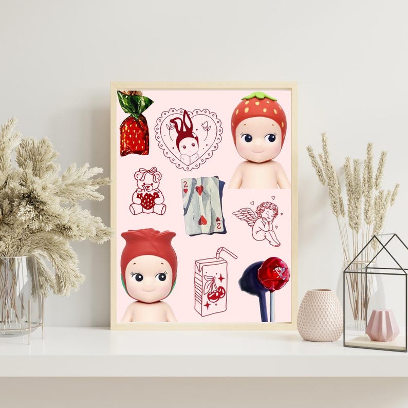 May include: A framed print featuring a collection of whimsical illustrations and dolls. The artwork includes a strawberry-themed doll, a red rose-shaped doll, a teddy bear, playing cards, a cherub, and a juice box with cherries. The background is a soft pink.
