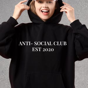 K&ouml;nnte beinhalten: Schwarzer Kapuzenpullover mit dem wei&szlig;en Text "ANTI- SOCIAL CLUB EST 2020". Der Hoodie hat eine Vordertasche und eine Kapuze. Unter der Kapuze wird eine wei&szlig;e Baseballkappe getragen.