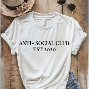 White tee on a wood background with the words anti-social club est. 2020