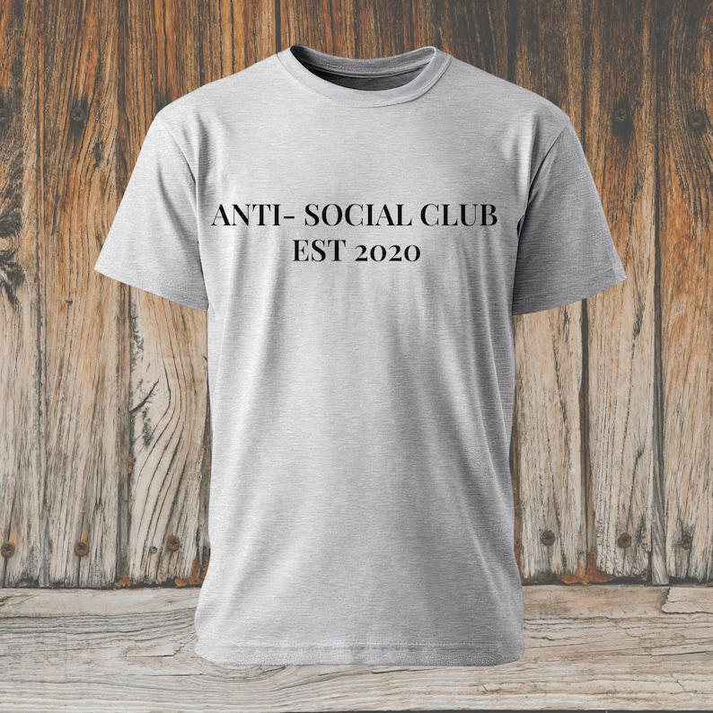 K&ouml;nnte beinhalten: Hellgraues T-Shirt mit dem schwarzen Text "ANTI- SOCIAL CLUB EST 2020". Das T-Shirt hat einen Rundhalsausschnitt und kurze &Auml;rmel. Der Hintergrund ist eine Holzoberfl&auml;che.