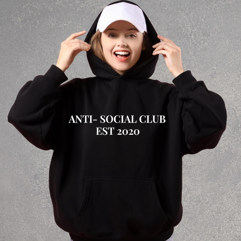 K&ouml;nnte beinhalten: Schwarzer Kapuzenpullover mit dem wei&szlig;en Text "ANTI- SOCIAL CLUB EST 2020". Der Hoodie hat eine Vordertasche und eine Kapuze. Unter der Kapuze wird eine wei&szlig;e Baseballkappe getragen.