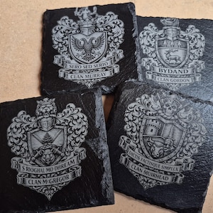 May include: Four square black slate coasters featuring laser-engraved Scottish clan crests. Each coaster displays a unique coat of arms with a shield, helmet, and banner. The text includes Clan Murray, Clan Gordon, Clan McGregor, and Clan Muirhead.