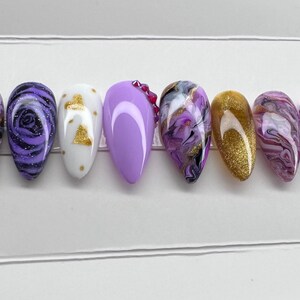May include: A collection of ten almond-shaped false nails. Designs include solid lavender, swirling purple and black patterns, gold glitter, and white with gold accents. Some nails are decorated with small, pink rhinestones. The nails are presented in a row.