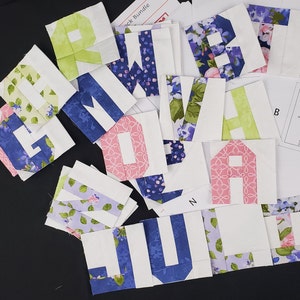 Pode incluir: Uma coleção de quadrados de tecido com letras do alfabeto impressas neles. As letras são de várias cores, incluindo azul, verde, rosa e branco. Os quadrados de tecido estão dispostos em um padrão aleatório sobre uma superfície preta.