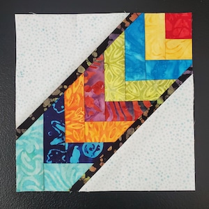 Puede incluir: Un bloque de colcha con un patrón diagonal de cuadrados de tela de colores. Los cuadrados están dispuestos en forma de zigzag y están bordeados por una tela negra con un patrón blanco. El fondo es una tela blanca con un patrón moteado azul claro.