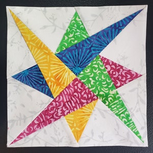 May include: A colorful patchwork quilt block featuring a star design with yellow, green, blue, and pink fabric pieces. The star is set against a white background with a subtle floral pattern.