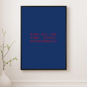 May include: A framed art print with a navy blue background and the red text: "KISS ALL THE TIME. DISCO, OCCASIONALLY." The print is in a black frame and is displayed on a white wall. A white vase with greenery is in the foreground.