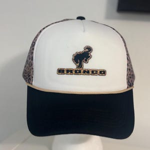 May include: A black and white trucker hat with a black brim and a white front panel. The front features a brown logo and the word "BRONCO" in gold lettering. The sides have a leopard print pattern.