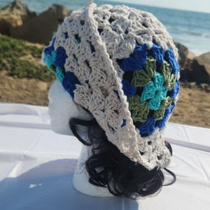 May include: A handmade crochet hat in a granny square pattern. The hat features a mix of colors, including cream, blue, green, and turquoise. The hat is displayed on a white mannequin head, with black hair peeking out from underneath.