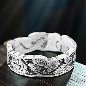 Può includere: Un anello color argento con un intricato design floreale e di foglie. L'anello è posto su una superficie di legno scuro, con i dettagli delle foglie e dei fiori chiaramente visibili. Il design dell'anello è intagliato nel metallo, creando un aspetto strutturato.