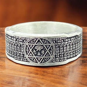 Puede incluir: Anillo plateado con un diseño detallado. Presenta un emblema central de una estrella de seis puntas con un ojo, rodeado de patrones y texto intrincados. Descansa sobre una superficie de madera.