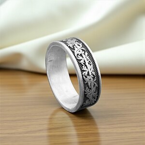 May include: A silver ring with an engraved floral pattern. The ring has a wide band with a detailed design of leaves and vines. The ring is displayed on a wooden surface with a white cloth in the background.