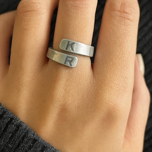 Pode incluir: Anel envolvente prateado com as letras "K" e "R" gravadas na banda. O anel é mostrado num dedo, com as letras claramente visíveis. Design simples e moderno, adequado para uso diário.