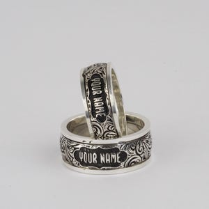 May include: Two silver rings with ornate scrollwork and the words "YOUR NAME" in a rectangular frame. One ring is stacked on top of the other, showcasing the design and text.
