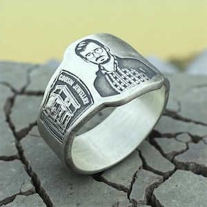 May include: A silver ring with an engraved portrait of a person wearing glasses and a checked shirt. The ring also features the text "COUSIN JEWELLRY" and a building image. The ring is shown on a cracked, grey surface.