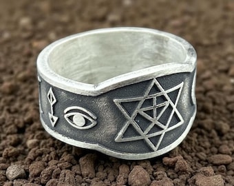 May include: A silver ring with a dark gray band featuring engraved symbols. The symbols include a six-pointed star, an eye, and other geometric shapes. The ring is set against a brown, textured background.