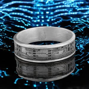 May include: A silver ring featuring a circuit board pattern engraved around the band. The ring reflects light, set against a dark backdrop with blue accents. A distinctive piece of jewellery.