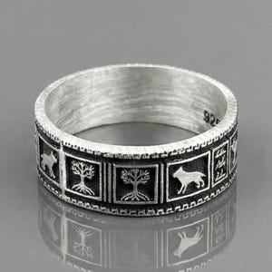 Peut inclure: Bague en argent avec un motif répétitif d'arbres et d'animaux dans des cadres rectangulaires. La bague a une bande texturée et est marquée "925". La bague est présentée sur une surface réfléchissante.