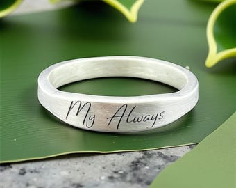 May include: A silver ring with the inscription "My Always" in black script. The ring is set against a green leaf, with a blurred background of green foliage. The ring has a brushed metal finish.