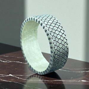 May include: A silver ring with a textured, scale-like pattern on the outer band. The ring has a wide band and a smooth, light-colored interior. The ring is displayed on a dark, marbled surface.