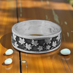May include: A silver ring with a detailed leaf pattern around the band. The ring is circular and rests on a wooden surface. The leaves are etched into the metal, creating a contrast with the silver background. Two small, white, oval stones are next to the ring.